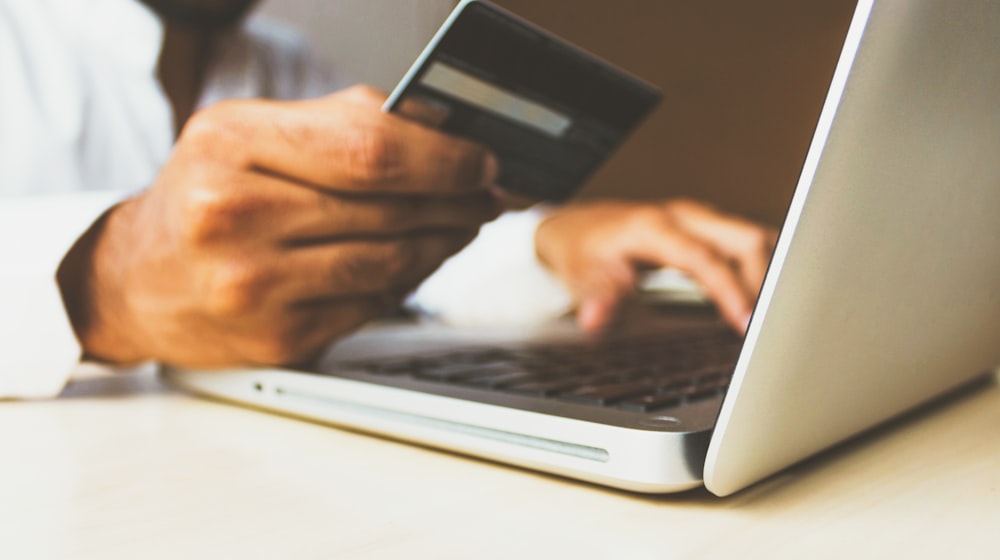 A customer holding a smartphone scans a PromptPay QR code displayed on a laptop checkout page in a small Thai boutique, while payment method icons for Visa, Mastercard, PayPal and Bitcoin line the bottom of the screen.